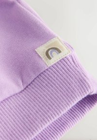 Indumento in cotone lilla con orlo a coste. Presenta un'etichetta quadrata con un semplice motivo arcobaleno in tonalità terrose. Vista ravvicinata della trama.