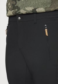 Pantalon noir en tissu lisse, avec une poche zippée à accent beige et un détail de bouton en métal. Pull à motif camouflé visible au-dessus.