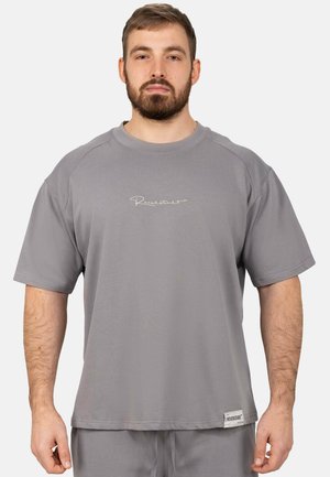 Bärtiger Mann trägt ein lockeres graues T-Shirt mit kurzen Ärmeln, kleinem weißen Text auf der Brust und einem Etikett nahe des Saums.