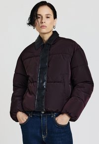 Daunenjacke in tiefem Burgunderrot mit einer schwarzen Kunstlederblende. Verfügt über einen Kragen und eine gesteppten Textur. Getragen über blauen Jeans.
