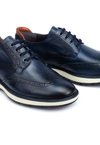 Chaussures en cuir bleu marine avec un design à bout ailé, détails perforés, bout rond et semelle blanche contrastante. Caractérisées par un système de laçage classique.