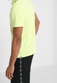 Limetkově zelená polo tričko s klasickým límečkem, krátkými rukávy a postranními ventily, v kombinaci s černými šortkami se vzorem opakovaných log.