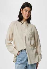 Jeune femme aux cheveux foncés portant une chemise beige ample boutonnée et un jean bleu clair, debout avec une main dans la poche sur un fond blanc.