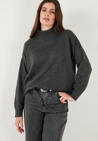 Grijze gebreide trui met een hoge, geribbelde kraag, losse pasvorm en verlaagde schouders. Gekleed met donkergrijze spijkerbroek en een zilveren riem.