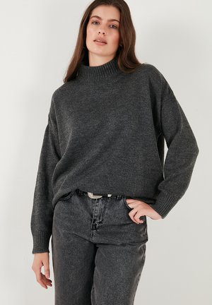 Gray knit sweater with a high, ribbed collar, loose fit, and drop shoulders. Paired with dark gray jeans and a silver belt.