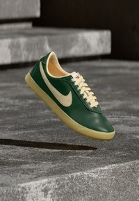 Πράσινο και μπεζ αθλητικό παπούτσι Nike, σε αναιρουμένη θέση πάνω από υφή σκαλοπατιών από σκυρόδεμα, παρουσιάζοντας πλευρική όψη με ορατά κορδόνια και λογότυπο.