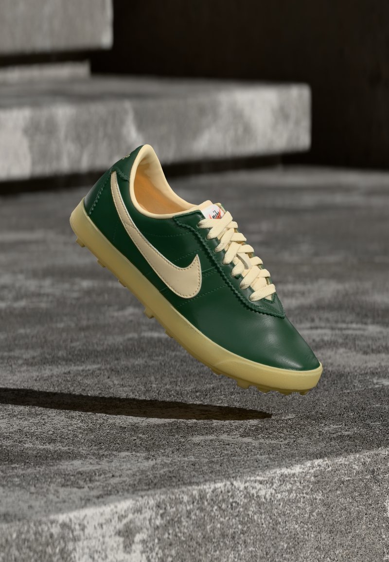 Πράσινο και μπεζ αθλητικό παπούτσι Nike, σε αναιρουμένη θέση πάνω από υφή σκαλοπατιών από σκυρόδεμα, παρουσιάζοντας πλευρική όψη με ορατά κορδόνια και λογότυπο.