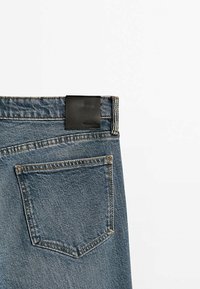Vue arrière d'un jean en denim bleu montrant la poche arrière droite et une étiquette de marque en cuir noir sur la ceinture, sur fond blanc.