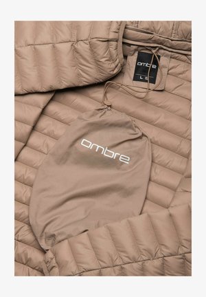 Cazadora de plumas marrón claro con textura de acolchado vertical y una bolsa de cuerda a juego. Presenta una etiqueta de logo en negro con texto blanco.