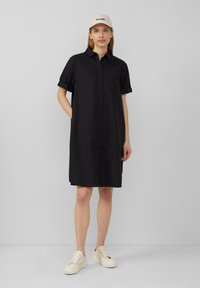 Abito camicia nero con maniche corte arrotolate, tasche laterali e vestibilità morbida, abbinato a sneakers di colore chiaro e un berretto.