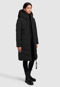 Vrouw in een lange zwarte pufferjas met een capuchon, zwarte leggings en zwarte enklaarsjes, staand tegen een effen grijze achtergrond.
