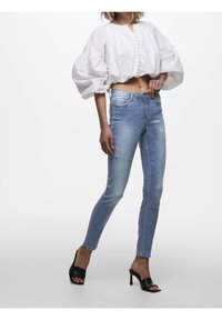 Blusa blanca corta con mangas abullonadas, cierre de botones en el frente y vaqueros skinny de lavado claro. Mules de tacón abierto negros completan el atuendo.