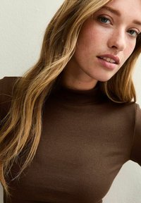 Top a collo alto marrone a coste con un design aderente. La modella ha capelli lunghi e lisci, lentiggini e uno sfondo neutro.