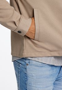 Beige Jacke mit einer Brusttasche und geknöpften Manschetten, über einem gestreiften Hemd getragen, kombiniert mit hellblauen, ausgewaschenen Jeans.