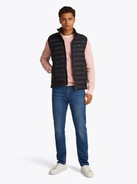 Colete puffer preto sobre um sweatshirt rosa, jeans azuis e ténis brancos. Apresenta fecho zip frontal e um design acolchoado vertical.