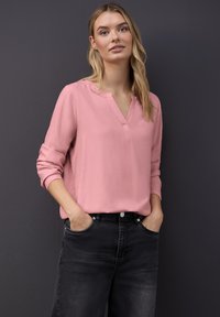 Rosa Bluse mit langen Ärmeln und V-Ausschnitt, aus glattem Stoff gefertigt, kombiniert mit dunklen Jeans. Die Hände sind in den Taschen.