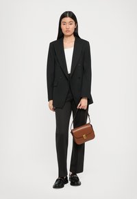 Svart dobbeltradet blazer over en hvit topp, kombinert med tettsittende brune bukser og svarte sko. Holder en brun skulderveske med gullfarget detaljer.