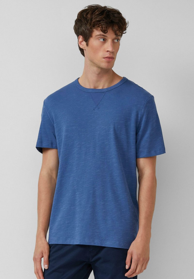 Jonge man met krullend haar, die een blauwe gemêleerde T-shirt met korte mouwen en donkere broeken draagt, kijkend naar zijn linkerzijde tegen een effen achtergrond.