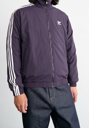Homme portant une veste de survêtement Adidas violette zippée avec des bandes blanches sur les manches et un jean bleu foncé devant un fond uni.