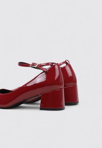Chaussures à talons blocs en cuir verni rouge avec bride de cheville. Présentent un bout arrondi et une texture lisse et brillante. Design minimaliste avec une couleur éclatante.