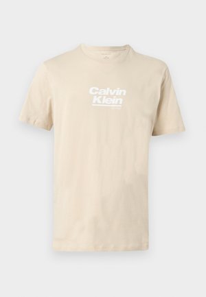 Beež lühikeste varrukatega ümara kaelusega T-särk, millel on valge "Calvin Klein est 1968" logo rinnal, esitatud lamedal, neutraalsel taustal.