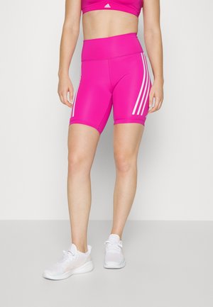 Žena v jasně růžových sportovních šortkách s vysokým pasem a bílými pruhy, bílých teniskách a sladěné růžové sportovní podprsence na jednobarevném pozadí.