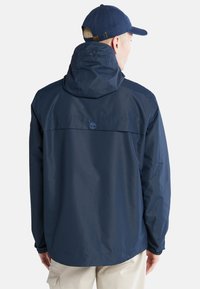 Giacca impermeabile navy con cappuccio, polsini elasticizzati e yoke posteriore. Presenta un piccolo logo vicino al collo. Texture liscia.