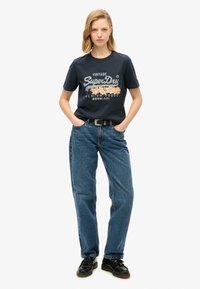 T-shirt in cotone navy con grafica "Vintage SuperDry" e stampa floreale, abbinato a jeans denim a gamba larga azzurri e scarpe nere.