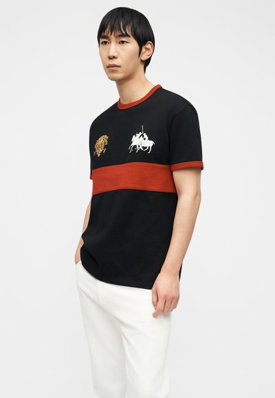 Νεαρός άνδρας που φοράει μαύρο και κόκκινο πόλο με κέντημα λευκού παίκτη πόλο με άλογο, σε συνδυασμό με λευκό παντελόνι σε απλό φόντο.