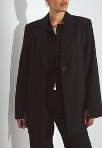Črn krojen blazer s strukturirano obliko, enojnim gumbom, izrazitimi reverji in dvema stranskima žepoma, oblečen čez temno srajco z gumbi.
