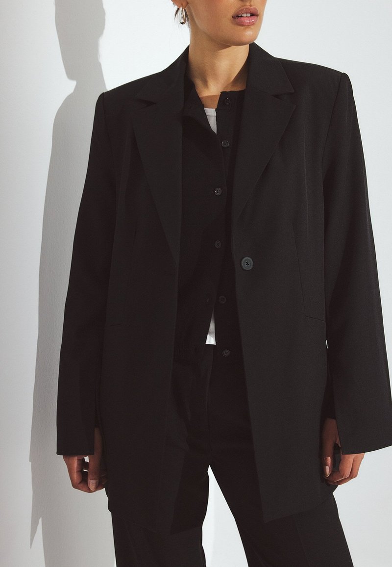 Črn krojen blazer s strukturirano obliko, enojnim gumbom, izrazitimi reverji in dvema stranskima žepoma, oblečen čez temno srajco z gumbi.