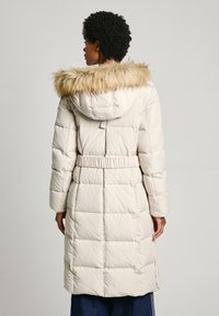 Vaddernad långkappa i ljusbeige med faux päls huva. Har en bälte i midjan och kviltad textur. Bakifrån syns vertikala sömmar och fickor.