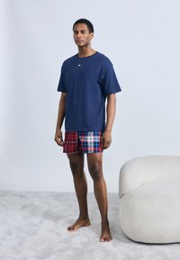 Μπλε βαμβακερό t-shirt με κοντά μανίκια, συνδυασμένο με κόκκινο και μπλε καρό σορτς. Το μοντέλο στέκεται ξυπόλητο σε ανοιχτογκρι χαλί κοντά σε μια μαλακή, κρεμ μπεζ πολυθρόνα.