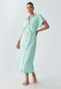 Mėtų žalios spalvos midi suknelė su diržu, trumpomis rankovėmis, mygtukų detalėmis ir dviem priekinėmis kišenėmis. Lygus audinys su laisvu siluetu.