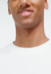 Man met een medium donkere huid, gekleed in een wit shirt met ronde hals, licht glimlachend, met wazige gezichtskenmerken en een lichte achtergrond.