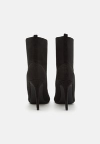 Mustat korkokengät nilkkurit, joissa on teksturoitu verkkokangas yläpinnassa ja sileät mokkapintaiset yksityiskohdat. Kengissä on terävä kärki ja hoikka stilettokorko.