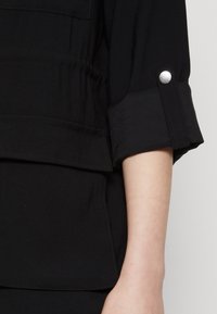 Camicia nera con colletto, dotata di polsini con bottone, dettagli plissettati e taschino frontale. Tessuto liscio con una leggera lucentezza.
