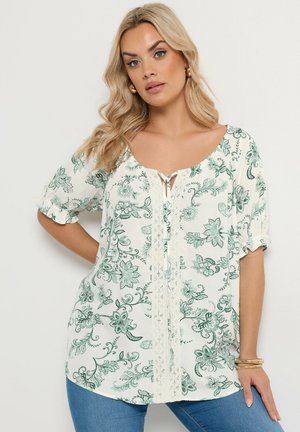 Kvinde med langt blondt hår iført en hvid bluse med grønt blomsterprint, blondedetaljer og blå jeans, stående mod en ensfarvet baggrund.