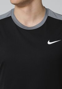 Sort atletisk t-shirt med grå krave og skulderdetaljer, med et lille hvidt Nike-logo på venstre bryst. Glat stofstruktur.