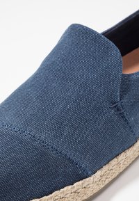 Chaussure à enfiler en toile marine avec une tige texturée, des détails cousus et une semelle naturelle en jute. Elle présente un bout arrondi et aucun élément métallique visible.