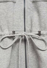 Haut de survêtement zippé gris clair avec taille à cordon, présentant un tissu texturé, des finitions en métal argenté et deux poches verticales à l'avant.