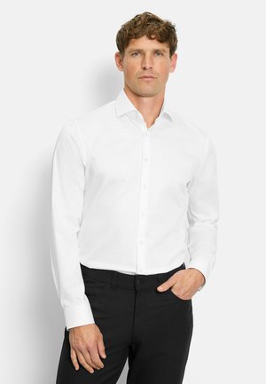 White button-up shirt with a spread collar, long sleeves, and a smooth texture. Paired with black trousers, hands resting casually.