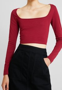 Burgundy pitkähihaine crop-top, jossa on neliönmuotoinen pääntie, valmistettu pehmeästä kankaasta. Se on yhdistetty korkeavyötäröiseen mustaan hameeseen, jossa on kaksi etutaskua.
