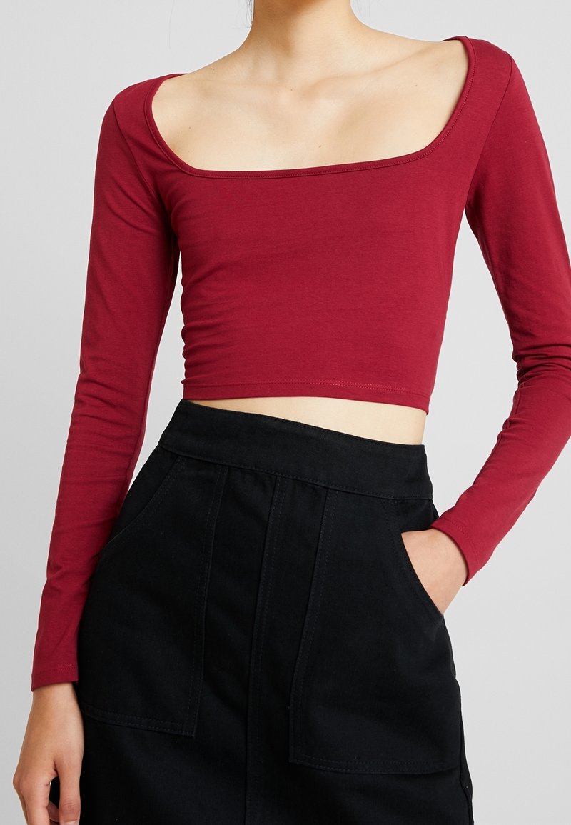 Burgundy pitkähihaine crop-top, jossa on neliönmuotoinen pääntie, valmistettu pehmeästä kankaasta. Se on yhdistetty korkeavyötäröiseen mustaan hameeseen, jossa on kaksi etutaskua.