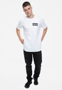 White cotton t-shirt with "oasis" printed in black on a rectangular label, paired with black pants and black footwear.