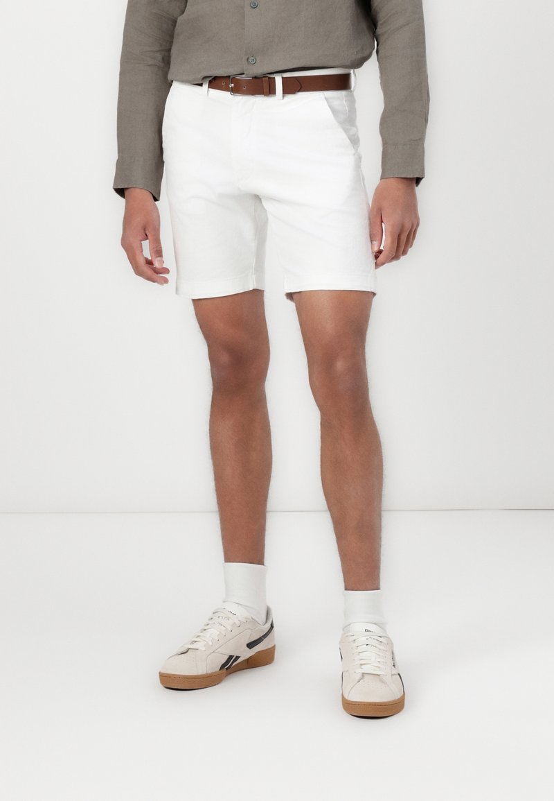 Shorts en coton blanc à coupe droite, stylisés avec une ceinture en cuir marron. Portés avec des baskets crème et des chaussettes blanches.