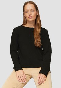 Chemise noire à manches longues texturée avec un col rond. Tissu doux et manches ajustées. Associée à un pantalon beige clair. Design simple et moderne.