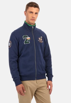 Felpa con zip di colore navy con colletto interno verde, decorata con ricami e toppe, tra cui un grande "2" verde e dettagli con la bandiera italiana.