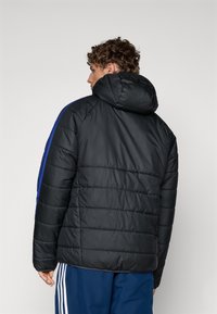 Svart boblejakke med hette, med blå side-striper, quiltet tekstur og elastiske mansjetter. Modellen står med ryggen til.