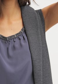 Cardigan grigio a maglia indossato sopra un top drappeggiato di un viola scuro con scollatura arricciata e rifiniture di perline decorate lungo il collo.